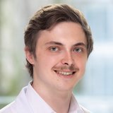 Porträt eines jungen Mannes im weißen Laborkittel, mit kurzem braunem Haar und Schnurrbart, lächelnd vor hellem, unscharfem Innenraum. ©Christian Houdek