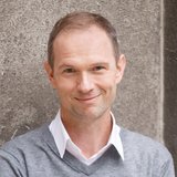Porträt eines lächelnden Mannes in grauem Pullover und weißem Hemd, vor einer Betonwand stehend ©derknopfdruecker.com